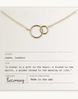 The Sisters Necklace by Becoming Jewelry features two intertwined gold circles on a delicate chain, presented on a card with an Isadora James quote about sisters. Made in the USA, this heartfelt gift beautifully symbolizes the bond between sisters.