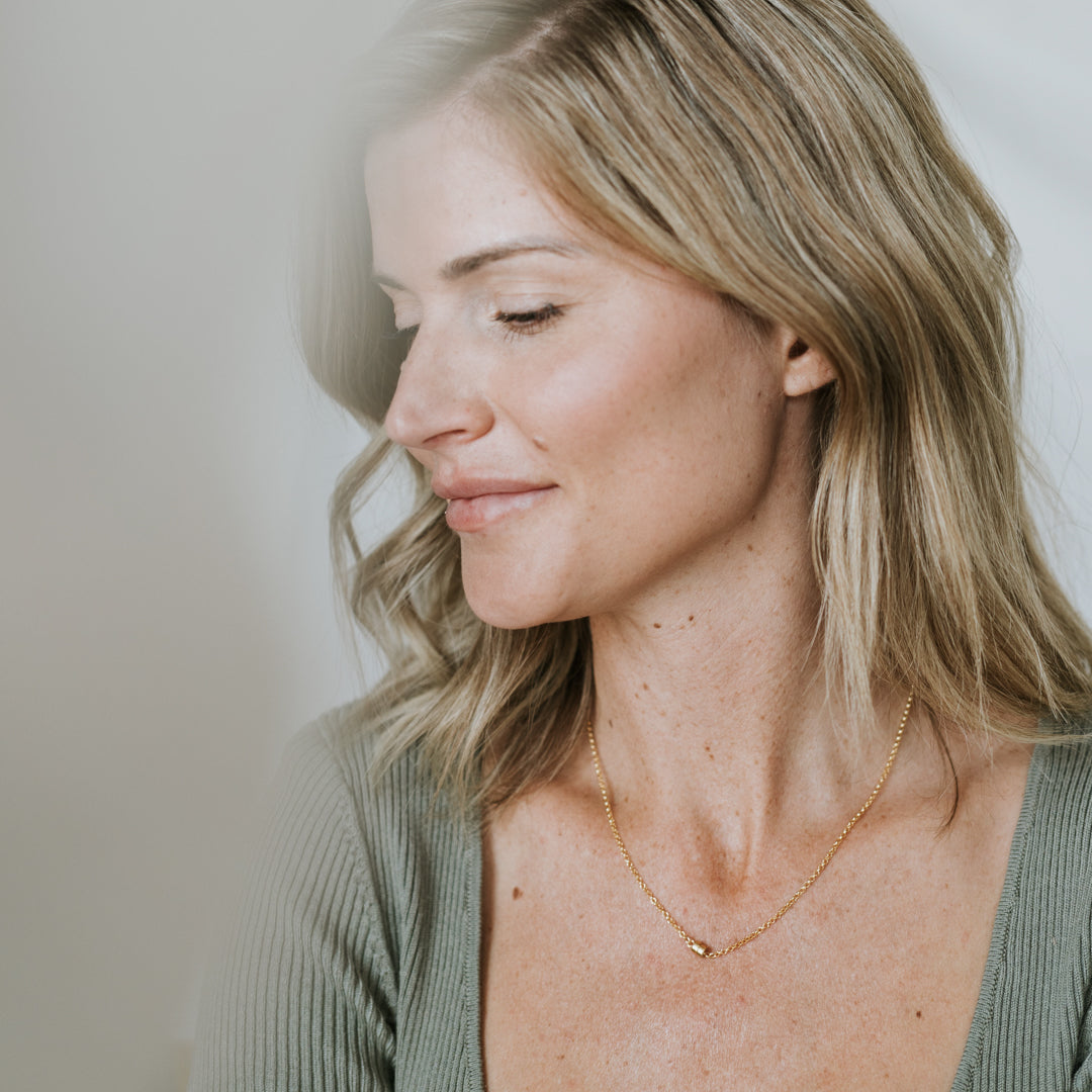 A woman with shoulder-length blonde hair, dressed in a green top, gazes downward with a serene expression, embodying a special connection to the Becoming Jewelry Stick Together Necklace of magnetic beads she wears.