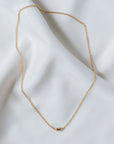 A charming Stick Together Necklace by Becoming Jewelry, showcasing a gold chain with a petite rectangular pendant, elegantly laid out on a white fabric background to represent a meaningful bond.