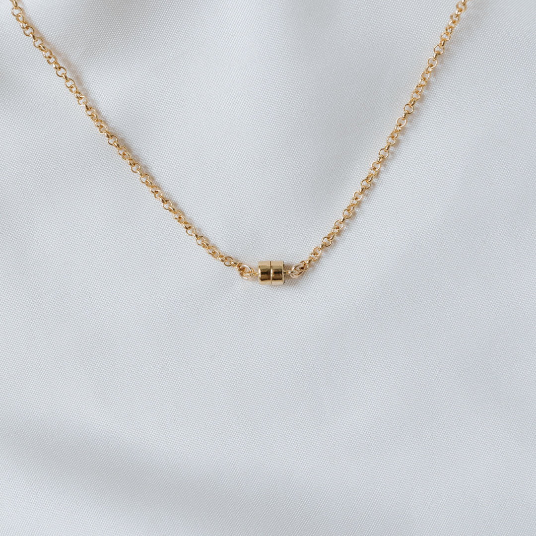 The Stick Together Necklace from Becoming Jewelry showcases a gold chain adorned with a cylindrical pendant, beautifully set against a white fabric background. This piece symbolizes meaningful connections, making it an ideal accessory for those who value cherished bonds.