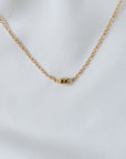 The Stick Together Necklace from Becoming Jewelry showcases a gold chain adorned with a cylindrical pendant, beautifully set against a white fabric background. This piece symbolizes meaningful connections, making it an ideal accessory for those who value cherished bonds.