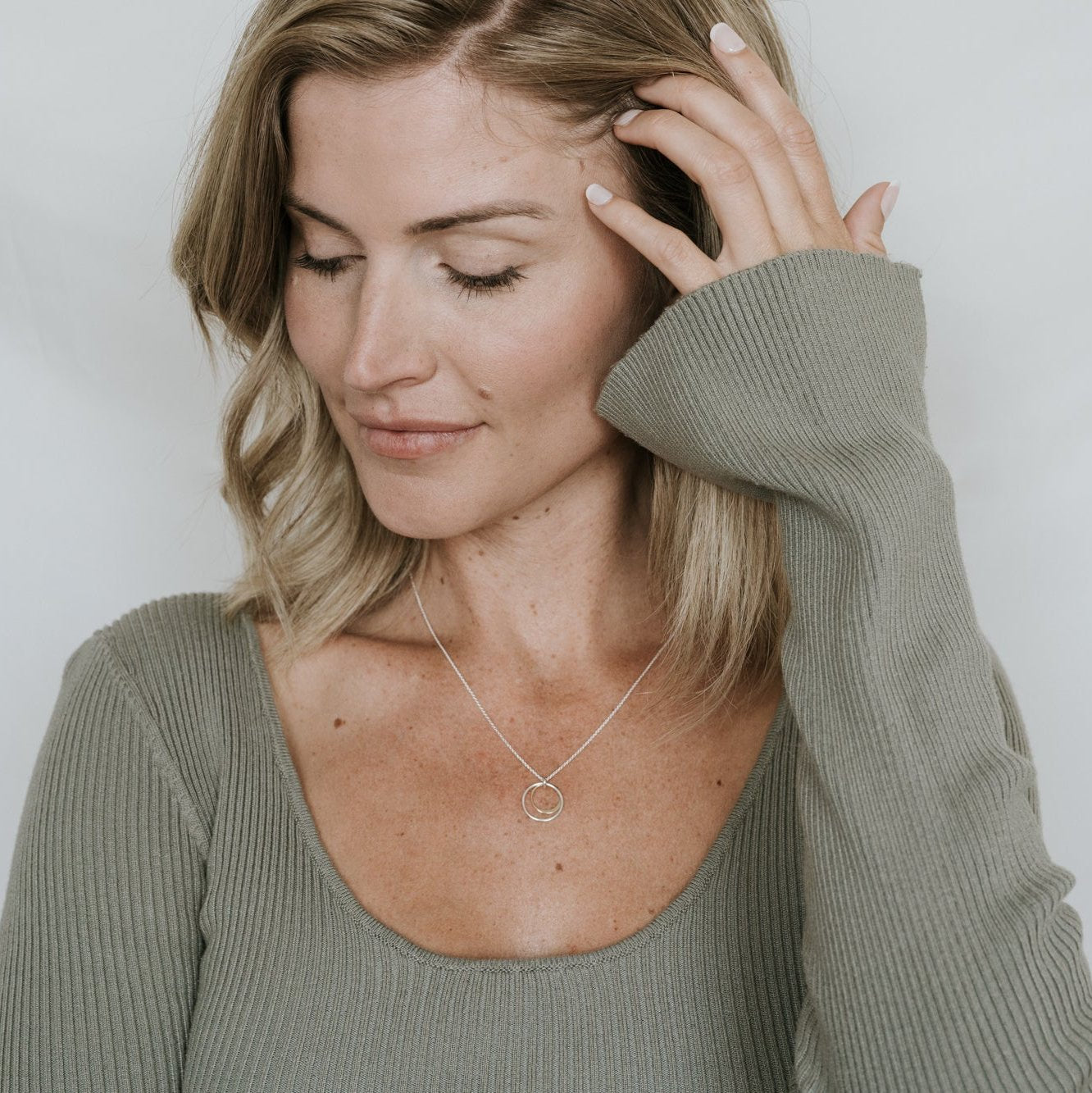 A woman with shoulder-length blonde hair wearing a green ribbed knit top and the "Surrounded By Love Necklace" by Becoming Jewelry as a meaningful reminder, looking down and touching her hair with her right hand.