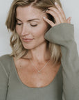 A woman with shoulder-length blonde hair wearing a green ribbed knit top and the "Surrounded By Love Necklace" by Becoming Jewelry as a meaningful reminder, looking down and touching her hair with her right hand.