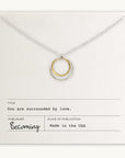 A simple necklace with interlocking rings displayed on a card that reads, "Surrounded By Love Necklace by Becoming Jewelry, Made in the USA," making it a meaningful reminder and a perfect gift to someone special.