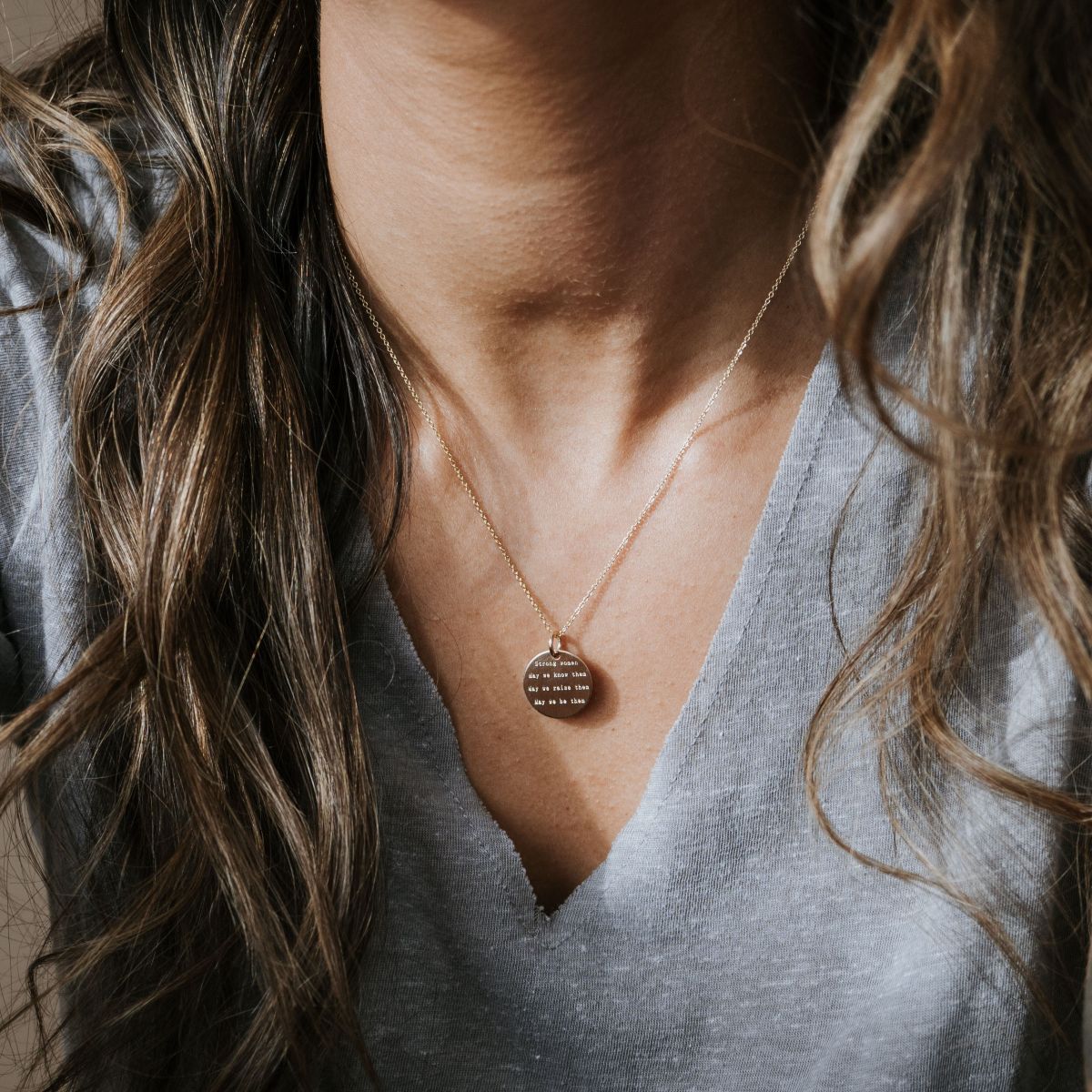Close-up of a person in a gray V-neck shirt wearing the Strong Women Necklace by Becoming Jewelry, featuring an engraved circular pendant. Their wavy brown hair partially covers their shoulders, making it perfect for everyday wear or as a thoughtful gift.