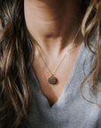 Close-up of a person in a gray V-neck shirt wearing the Strong Women Necklace by Becoming Jewelry, featuring an engraved circular pendant. Their wavy brown hair partially covers their shoulders, making it perfect for everyday wear or as a thoughtful gift.