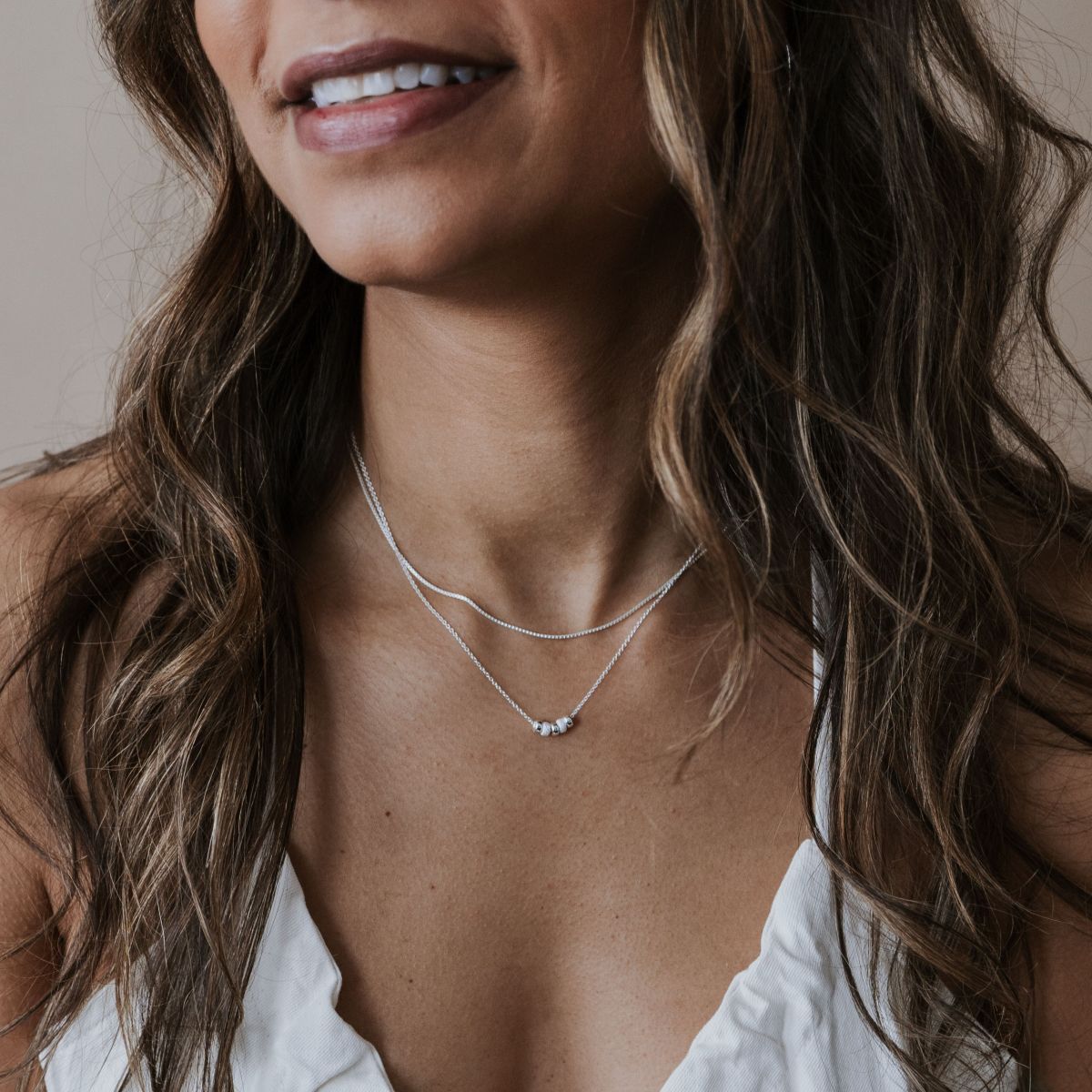 A woman in a white top wears the My Wish For You Necklace from Becoming Jewelry, featuring two silver chains with tiny beads and a small pendant. Her wavy hair flows over her shoulders, enhancing her gentle smile.