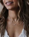 A woman in a white top wears the My Wish For You Necklace from Becoming Jewelry, featuring two silver chains with tiny beads and a small pendant. Her wavy hair flows over her shoulders, enhancing her gentle smile.
