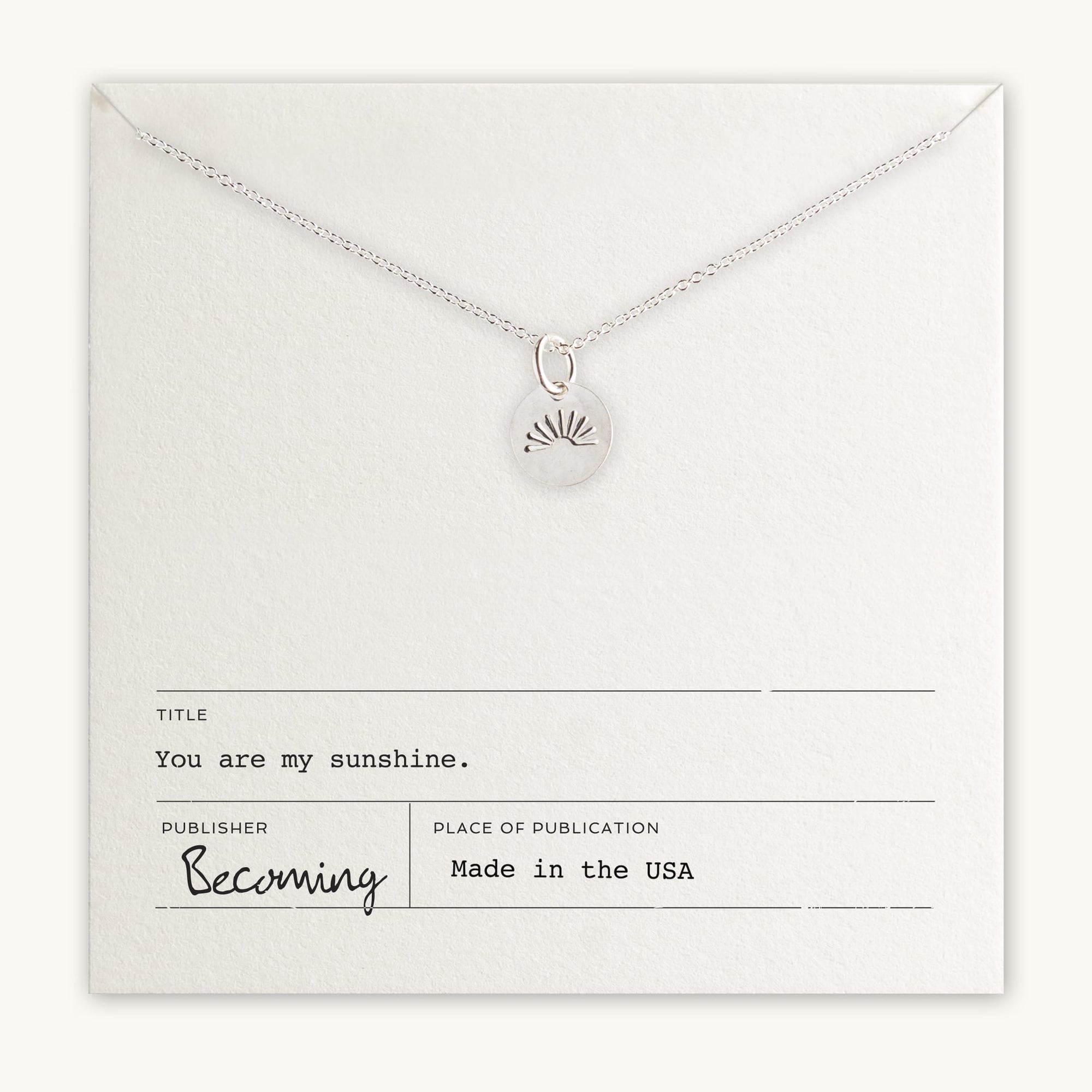 The bestselling You Are My Sunshine Necklace from Becoming Jewelry features a radiant sun charm and is presented on a cream card with the messages "You are my sunshine" and "Becoming, Made in the USA." This silver gift eloquently conveys love and warmth.