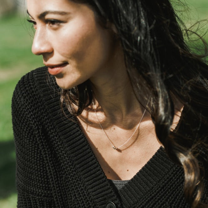 A woman with long dark hair, dressed in a black knit sweater and the Three Things Necklace from Becoming Jewelry, gazing to the side in an outdoor setting, exuding grace.