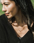 A woman with dark hair wearing a black top and a delicate Becoming Jewelry Three Things Necklace, gazing to the side outdoors.