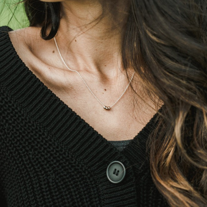 A woman wearing a black knitted cardigan and the Becoming Jewelry "Three Things Necklace" with a simple silver pendant, exuding grace and quiet encouragement.