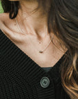 Close-up of a woman wearing a delicate Becoming Jewelry Three Things Necklace and a black knitted sweater with a button visible.
