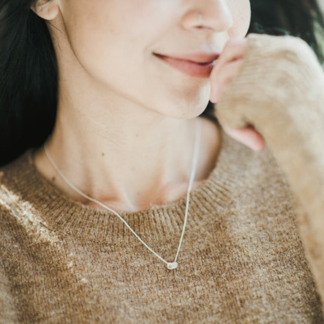A woman smiling gently with her chin resting on her hand, wearing a Becoming Jewelry Blessings Necklace with a small pendant.