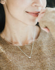 A woman smiling gently with her chin resting on her hand, wearing a Becoming Jewelry Blessings Necklace with a small pendant.