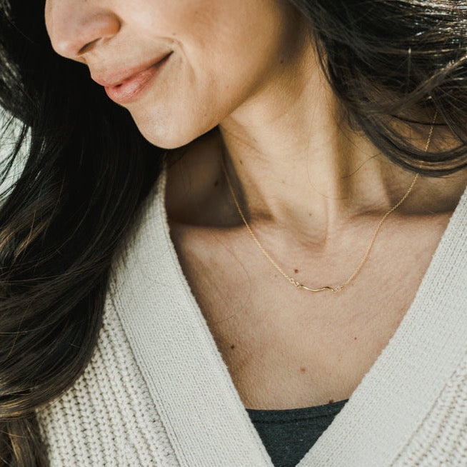 A woman wearing a light knit sweater and Becoming Jewelry's Path Necklace, a delicate gold piece that serves as a gentle reminder of their meaningful gift, with long dark hair cascading over her shoulder.