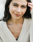 A woman with long dark hair wears a beige knit cardigan and a necklace adorned with intertwined circles. She smiles slightly while touching her hair, the Sisters Necklace—a gift from her sister by Becoming Jewelry—gleaming softly in the light.