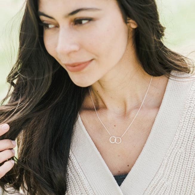 A woman wearing a white cardigan and a Becoming Jewelry "Joined for Life" necklace with intertwined circles stands outside, looking to the side while touching her hair.