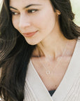 A woman wearing a white cardigan and a Becoming Jewelry "Joined for Life" necklace with intertwined circles stands outside, looking to the side while touching her hair.