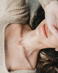 A woman wearing a Friendship Circles Necklace by Becoming Jewelry lies on her back, dressed in a beige knit sweater, with her right hand partially covering her face.