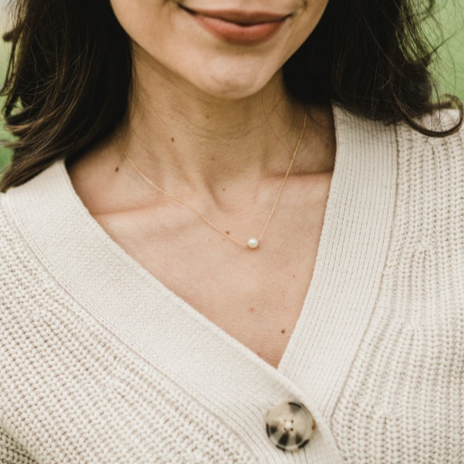 A woman wearing a beige knit cardigan with a single button fastened, and an elegant Simple Pleasures Pearl Necklace from Becoming Jewelry, adorned with a single pearl pendant, exuding timeless beauty.