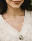 A woman wearing a beige knit cardigan with a single button fastened, and an elegant Simple Pleasures Pearl Necklace from Becoming Jewelry, adorned with a single pearl pendant, exuding timeless beauty.
