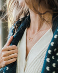Close-up of a person wearing a white knitted top and a navy blue patterned cardigan, showcasing the Becoming Jewelry Mother Necklace with interlocking rings symbolizing mother's love, and a gold ring on their finger.