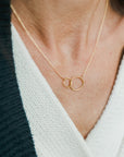 A woman wearing a white v-neck garment and a gold Becoming Jewelry mother necklace with two interlocking circles.