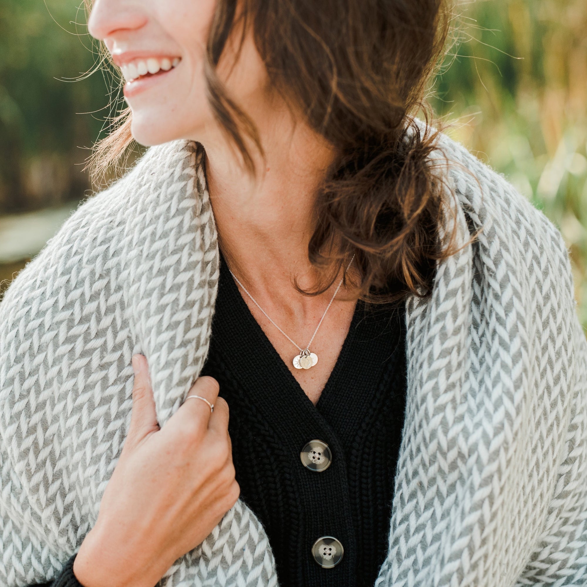 A smiling woman wears a black buttoned cardigan and a gray patterned scarf. She proudly displays the "Sun Moon and Stars Necklace" by Becoming Jewelry, featuring circular pendants with a delicate moon charm, and has a simple ring on her finger. The background is outdoors and slightly blurred.
