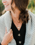 A smiling woman wears a black buttoned cardigan and a gray patterned scarf. She proudly displays the "Sun Moon and Stars Necklace" by Becoming Jewelry, featuring circular pendants with a delicate moon charm, and has a simple ring on her finger. The background is outdoors and slightly blurred.