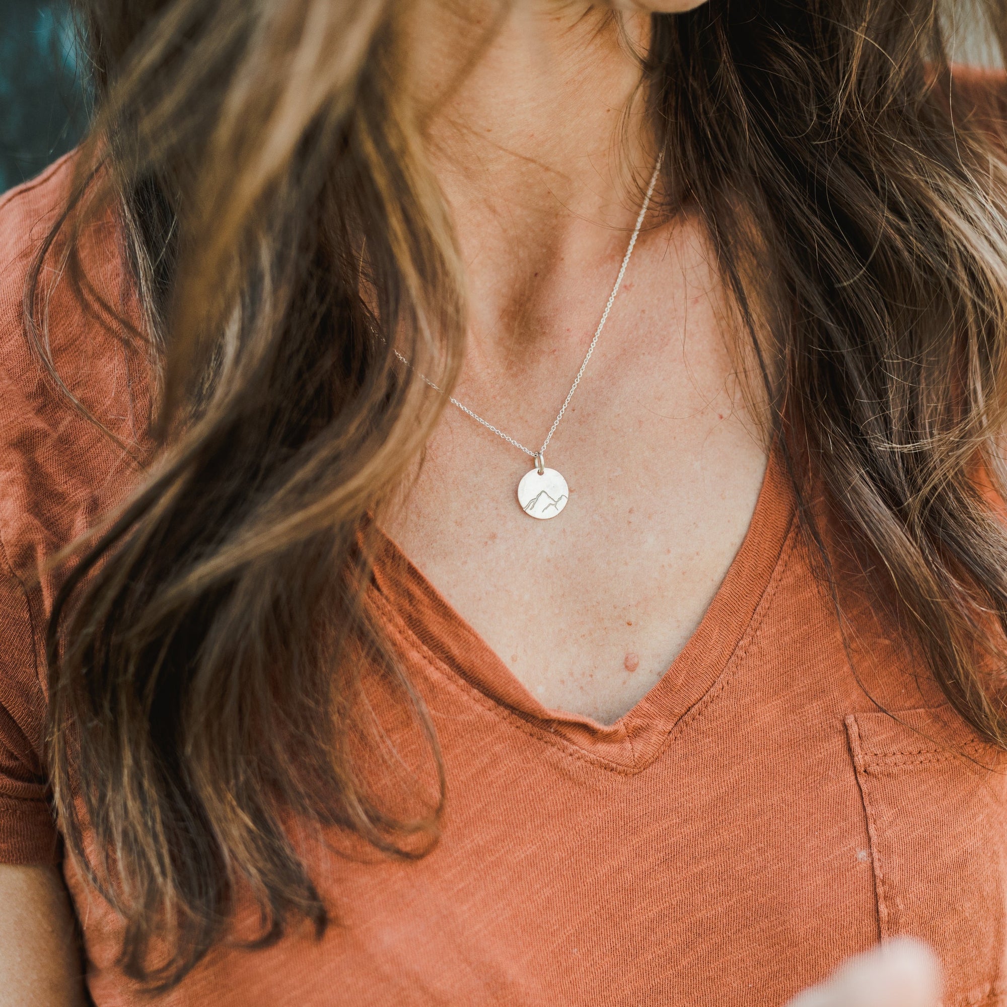 A person in an orange V-neck shirt is wearing the "Mountains Are Calling Necklace" by Becoming Jewelry, showcasing a round pendant with a small mountain charm, ready for their next adventure.