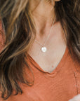 A person in an orange V-neck shirt is wearing the "Mountains Are Calling Necklace" by Becoming Jewelry, showcasing a round pendant with a small mountain charm, ready for their next adventure.