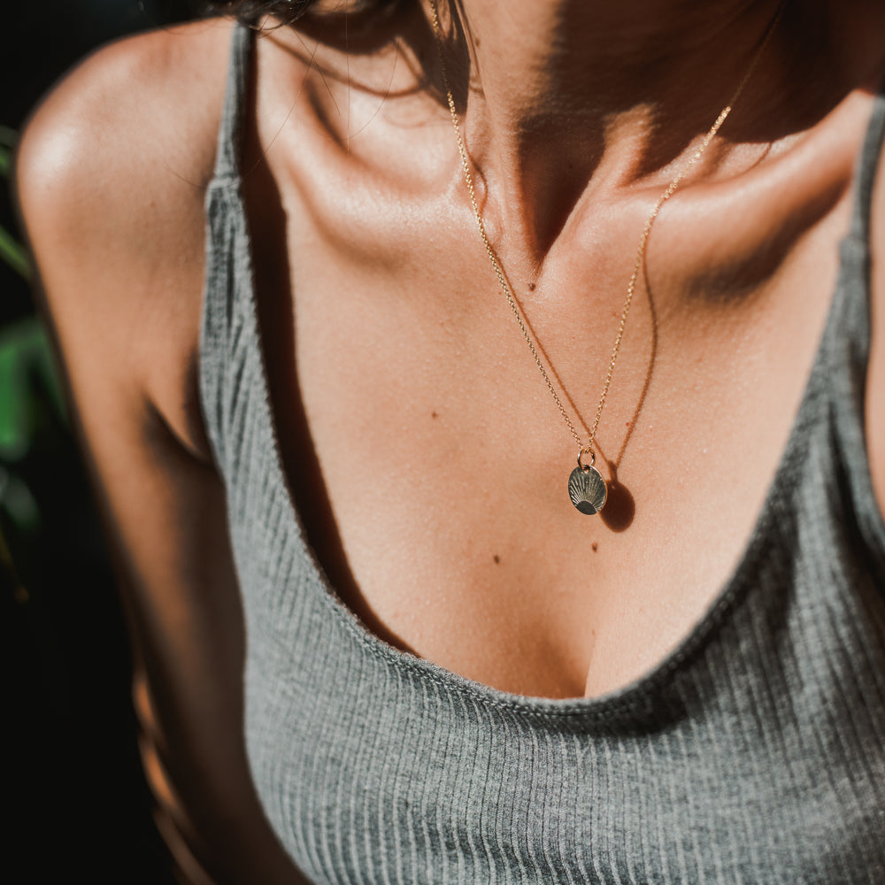 Close-up of a woman wearing the Irish Blessing Necklace by Becoming Jewelry, featuring a small shell pendant, and a grey ribbed tank top. The background is dark with some blurred green elements, evoking the spirit of new adventure.