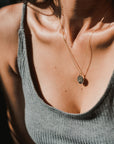 Close-up of a woman wearing the Irish Blessing Necklace by Becoming Jewelry, featuring a small shell pendant, and a grey ribbed tank top. The background is dark with some blurred green elements, evoking the spirit of new adventure.