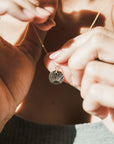 A person holds a Becoming Jewelry Irish Blessing Necklace, adorned with a circular pendant featuring a rays design, captured in a close-up view of their hands and part of their upper torso.