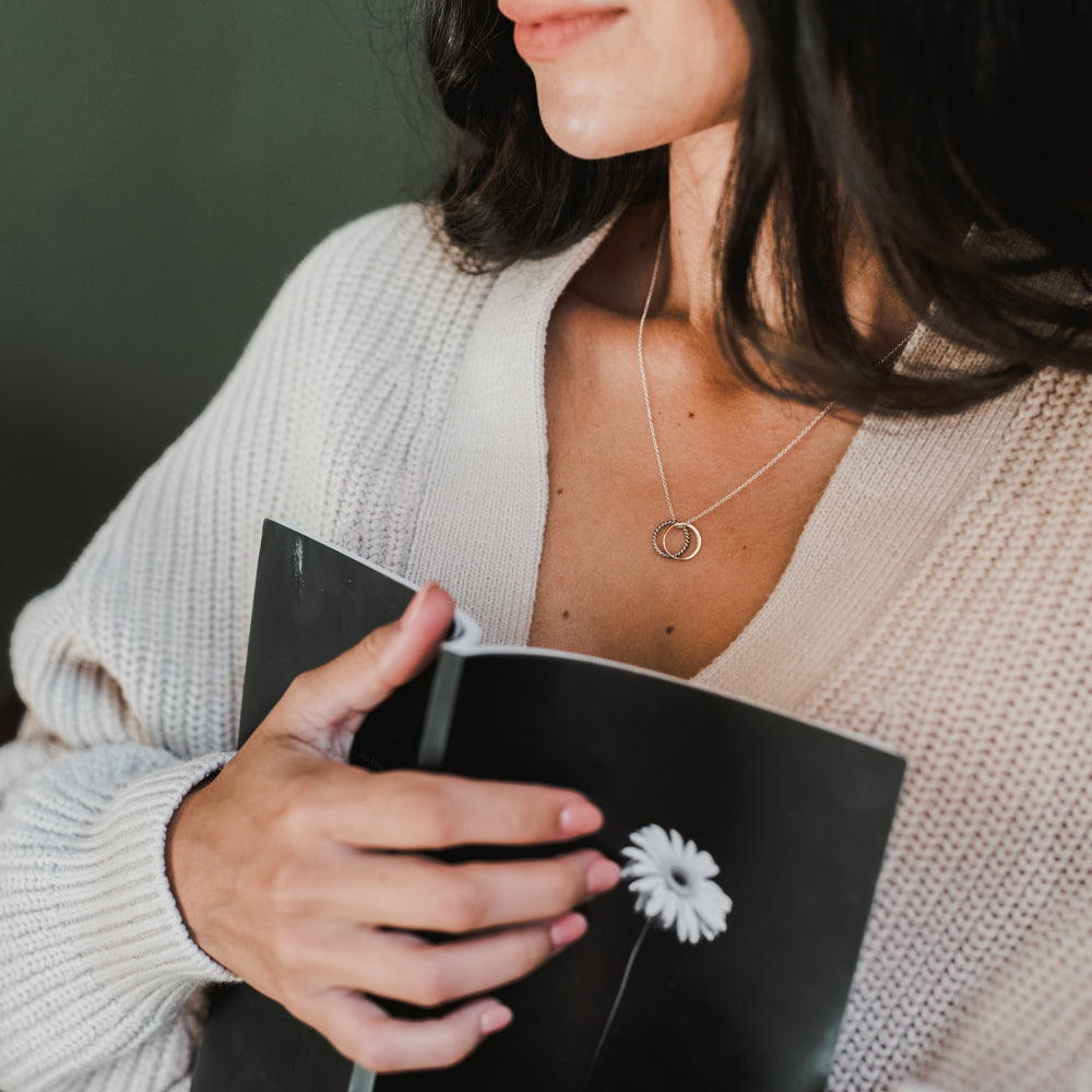 A person holds a black notebook adorned with a white flower on the cover, wearing a beige knit cardigan and the True Friends Necklace by Becoming Jewelry, which features two mixed metal intertwined rings.
