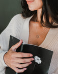 A person holds a black notebook adorned with a white flower on the cover, wearing a beige knit cardigan and the True Friends Necklace by Becoming Jewelry, which features two mixed metal intertwined rings.