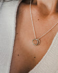 A person wearing a knitted sweater and the True Friends Necklace by Becoming Jewelry, featuring two interlocking rings.
