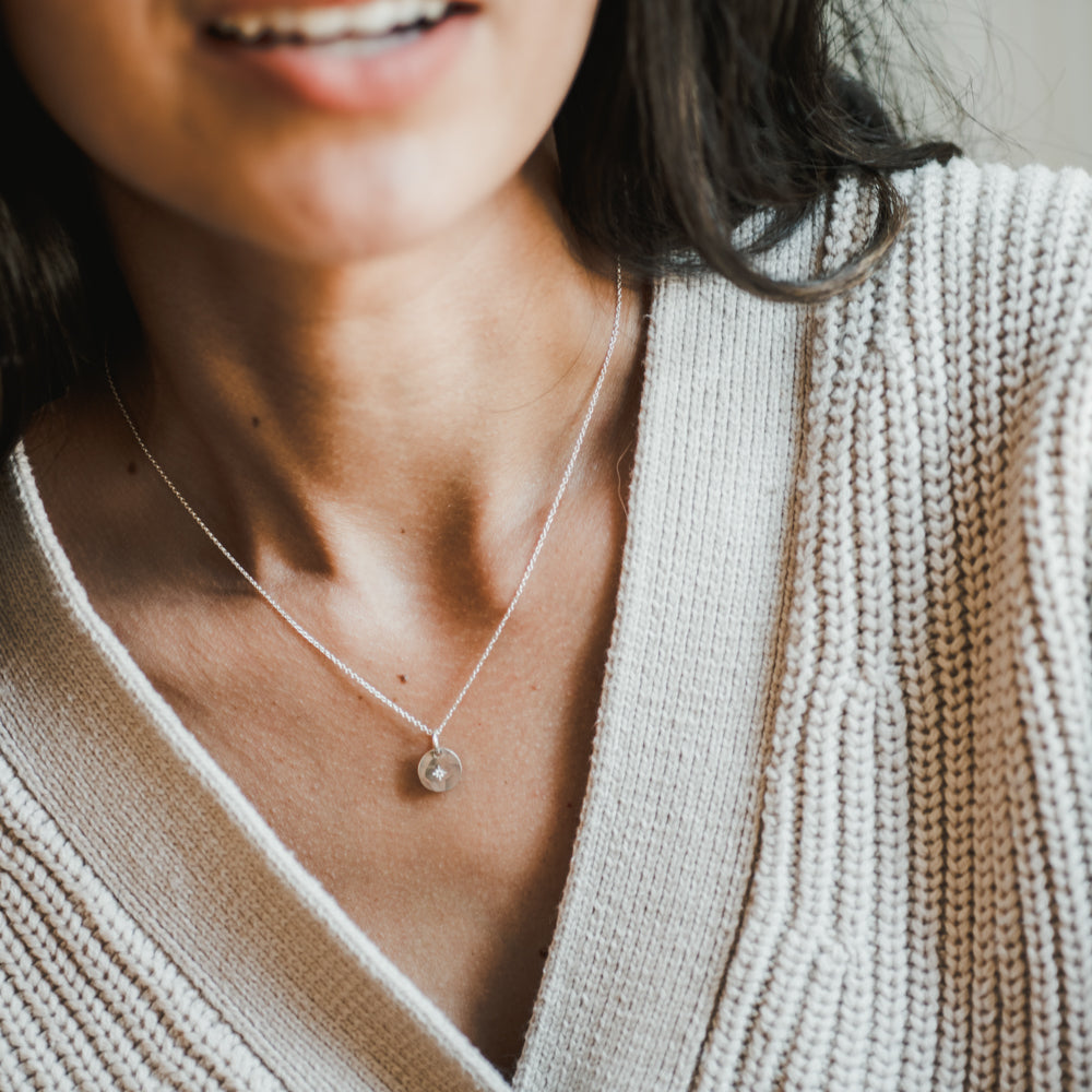 A close-up of a person wearing a light-colored knitted sweater and the Light Within Necklace by Becoming Jewelry, which features a round pendant adorned with a tiny starburst. The person's face is partially visible, blending seamlessly with the light within the delicate design.