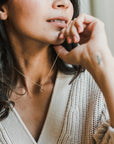 Close-up of a person wearing a light sweater, resting their chin on their hand, and adorned with the delicate Light Within Necklace from Becoming Jewelry, featuring a tiny starburst pendant. The person has dark hair and a light skin tone.