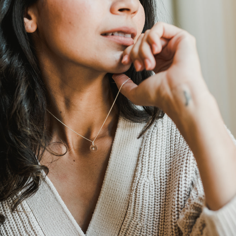 Woman in a beige sweater with a gold filled, delicate Light Within Necklace by Becoming Jewelry featuring a starlight charm, contemplatively touching her chin.