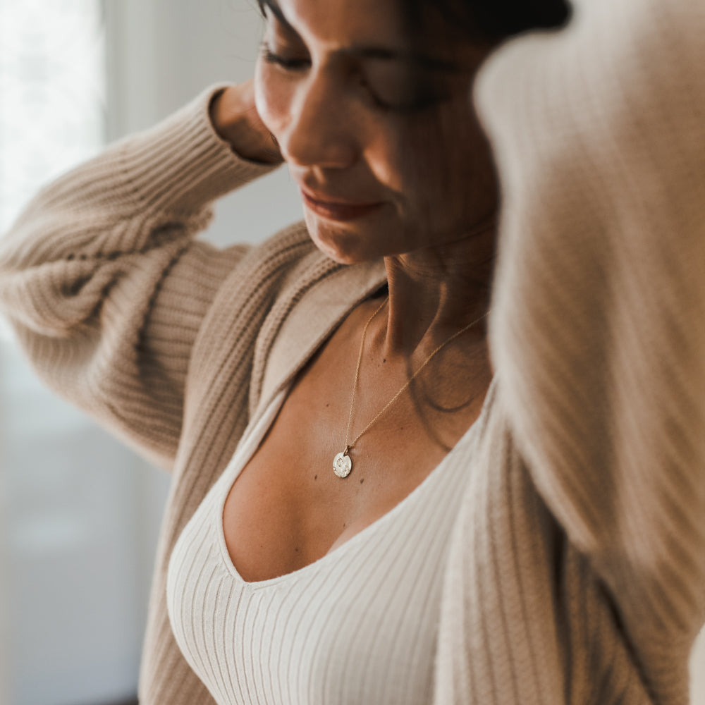 A person wearing a light-colored ribbed top and a beige cardigan adjusts their hair. They have Becoming Jewelry's Blossom Necklace on and are looking down with a soft smile, reminiscent of blossoming flowers.