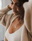 A person wearing a light-colored ribbed top and a beige cardigan adjusts their hair. They have Becoming Jewelry's Blossom Necklace on and are looking down with a soft smile, reminiscent of blossoming flowers.