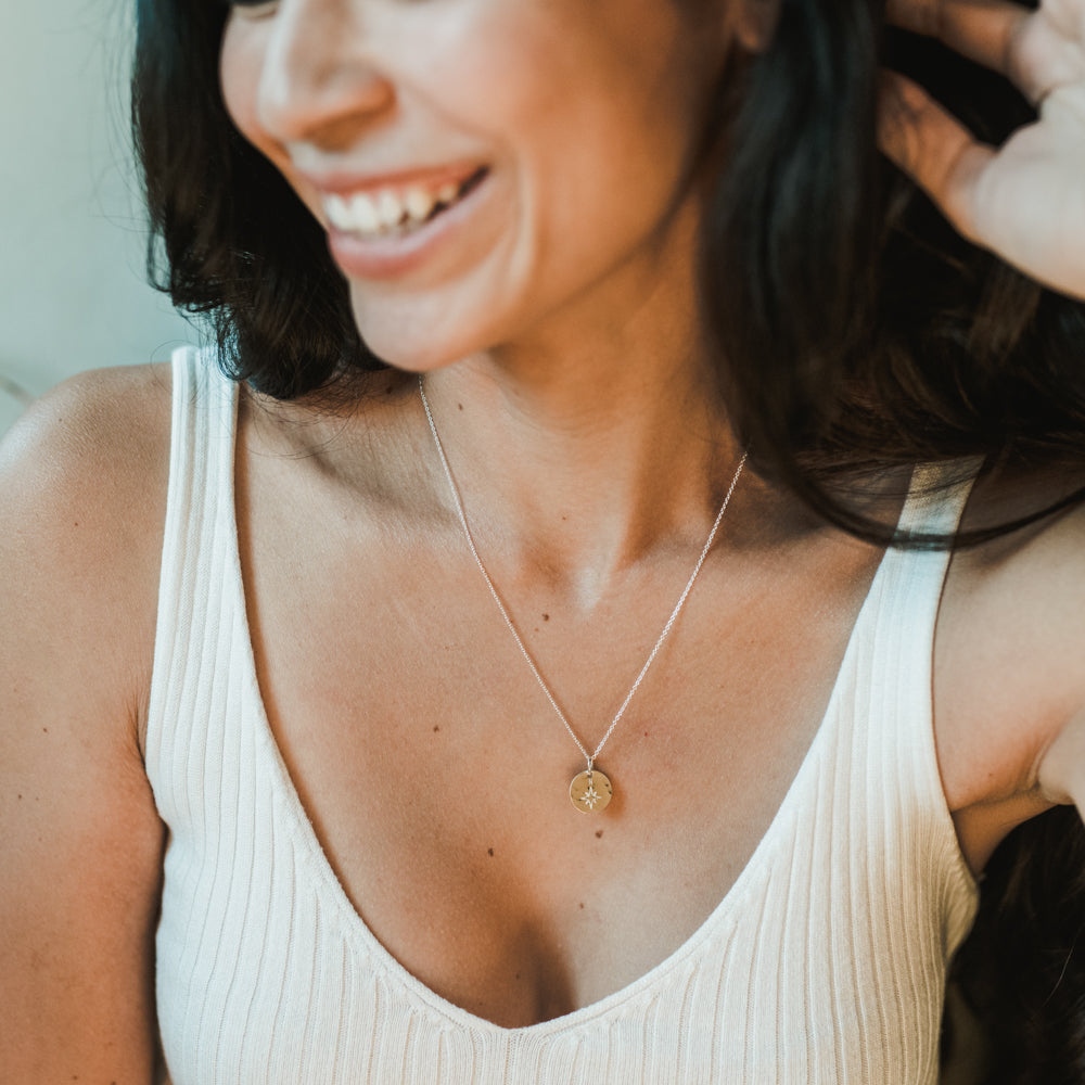 A woman wearing a white sleeveless top smiles, showcasing the North Star Necklace by Becoming Jewelry—an exquisite piece perfect as a graduation gift.