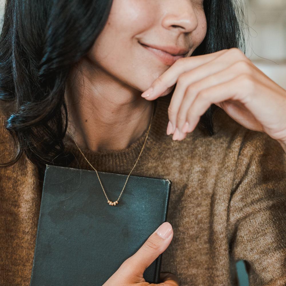 A person with long dark hair wearing a brown sweater holds a closed book close to their chest, smiling softly. They wear the "My Wish For You Necklace" by Becoming Jewelry, featuring a small gold pendant inscribed with the heartfelt message "Live in the sunshine," inspired by a Ralph Waldo Emerson quote.