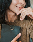 A person with long dark hair wearing a brown sweater holds a closed book close to their chest, smiling softly. They wear the "My Wish For You Necklace" by Becoming Jewelry, featuring a small gold pendant inscribed with the heartfelt message "Live in the sunshine," inspired by a Ralph Waldo Emerson quote.