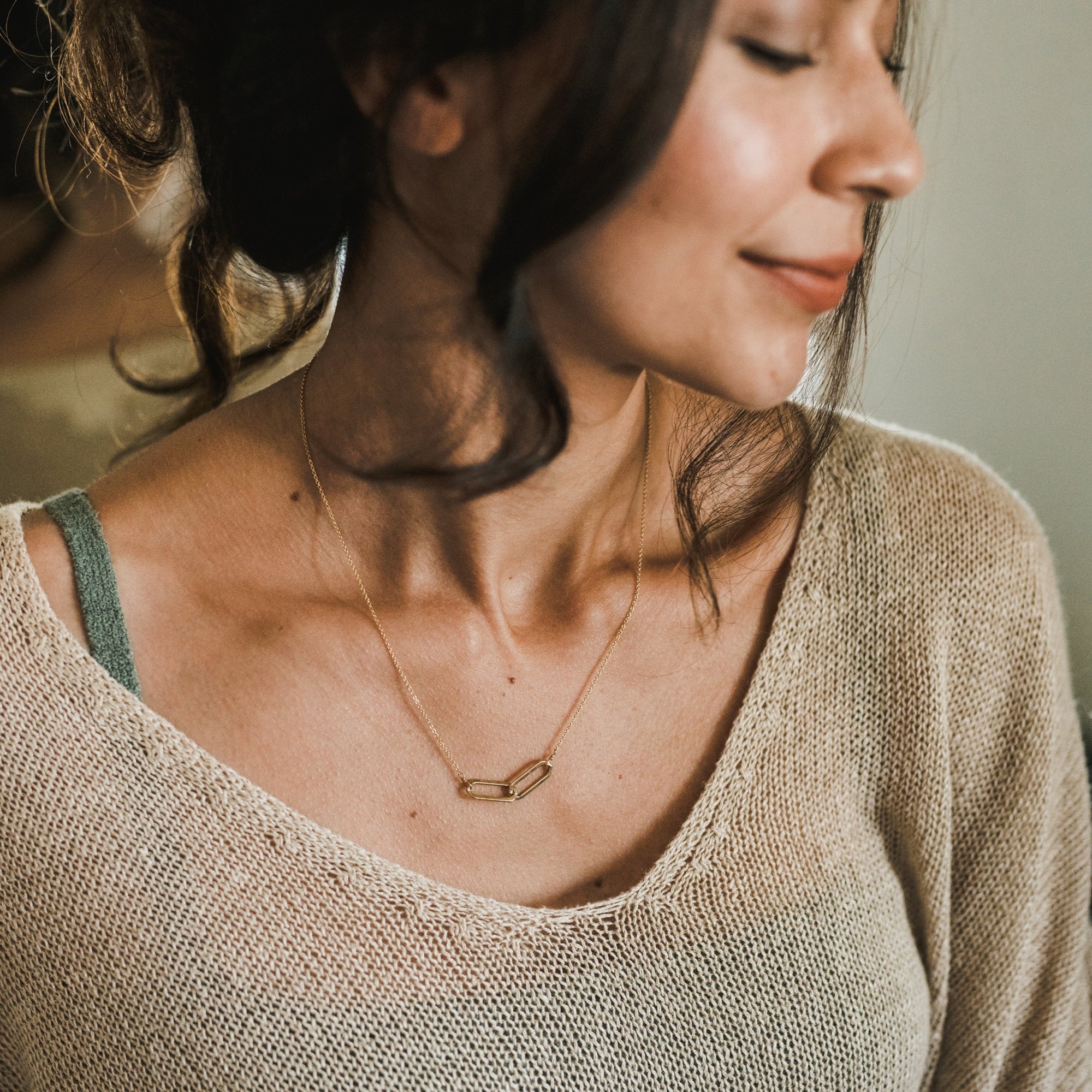 A person with dark hair is wearing a beige knit top and a delicate gold Linked Together Necklace from Becoming Jewelry, featuring a rectangular pendant. They are looking down, smiling softly, perhaps reflecting on the special connection it signifies.