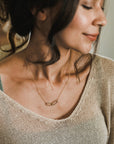A person with dark hair is wearing a beige knit top and a delicate gold Linked Together Necklace from Becoming Jewelry, featuring a rectangular pendant. They are looking down, smiling softly, perhaps reflecting on the special connection it signifies.