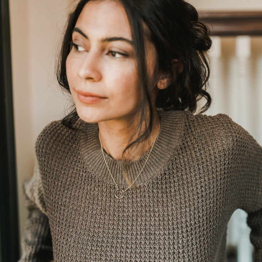 Woman in a knitted sweater adorned with a Becoming Jewelry Karma Necklace, looking away from the camera.
