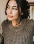 Woman in a knitted sweater adorned with a Becoming Jewelry Karma Necklace, looking away from the camera.